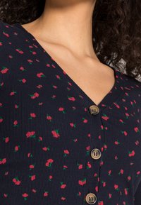 Marinefarbener Cardigan mit Knopfleiste und geripptem Muster, verziert mit einem Kirschmuster in Rot und Grün, sowie fünf runden beigen Knöpfen auf der Vorderseite.