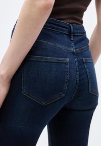 Κοντινό πλάνο ενός ατόμου που φοράει σκούρα μπλε τζιν και μαύρο τοπ, δείχνοντας την πίσω τσέπη και τις λεπτομέρειες της ραφής.