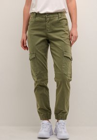 Pantalons cargo vert olive avec poches, coupe fuselée, poignets élastiques et coutures contrastées, associés à des chaussures de sport blanches.