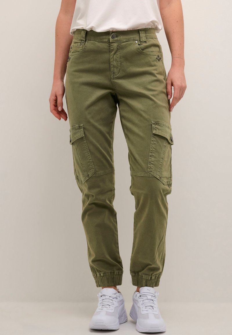 Pantalons cargo vert olive avec poches, coupe fuselée, poignets élastiques et coutures contrastées, associés à des chaussures de sport blanches.