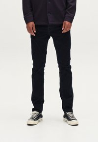 Tamsiai mėlynos džinsinės kelnės, kūno prigludusio silueto, dėvimos su juodai baltais drobės sportbačiais; fone - paprasta balta spalva, kad sukurtų kontrastą.