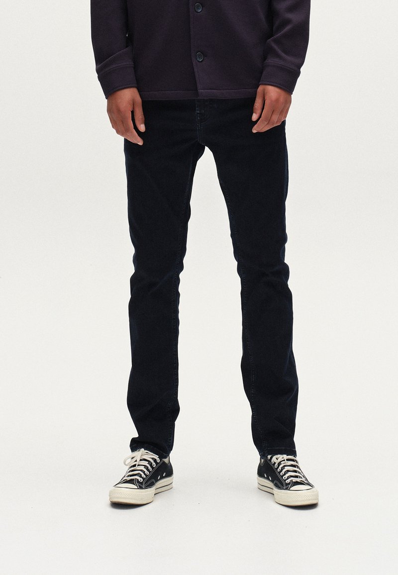 Tamsiai mėlynos džinsinės kelnės, kūno prigludusio silueto, dėvimos su juodai baltais drobės sportbačiais; fone - paprasta balta spalva, kad sukurtų kontrastą.