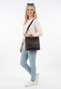 Donna sorridente, indossa un maglione rosa chiaro, jeans azzurri, sneaker bianche, orologio viola, borsa a tracolla nera e occhiali da sole sulla testa.