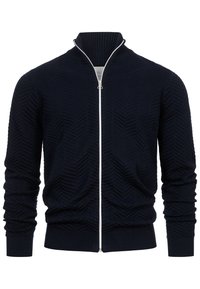 Marineblå zip-up cardigan laget av teksturert stoff med et sikksakkmønster, ribbestrikkede mansjetter og en høy krage med en kontrasterende hvit glidelås.