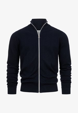 Marineblå zip-up cardigan laget av teksturert stoff med et sikksakkmønster, ribbestrikkede mansjetter og en høy krage med en kontrasterende hvit glidelås.