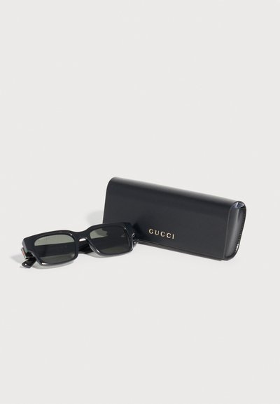 Μαύρα ορθογώνια γυαλιά ηλίου Gucci δίπλα σε μαύρη θήκη με χρυσό λογότυπο Gucci σε λευκό φόντο.