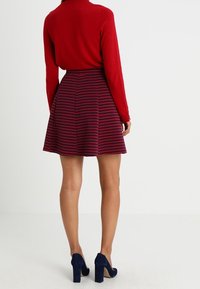 Maglione rosso abbinato a una gonna viola a fantasia, caratterizzata da righe orizzontali. L'outfit è completato da tacchi alti blu.