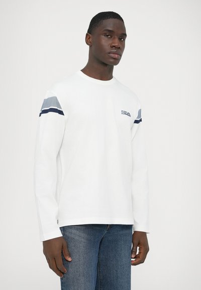 Λευκό πουκάμισο με μακριά μανίκια, με μπλε και γκρι ριγέ λεπτομέρειες στους ώμους και μικρό λογότυπο στο στήθος. Το ύφασμα φαίνεται μαλακό και απαλό.