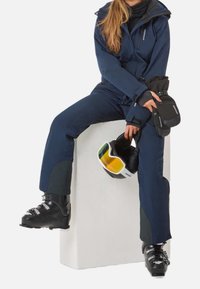 Personne assise portant une combinaison de ski bleu marine et des chaussures de ski noires, tenant des gants noirs et un masque de ski blanc avec des verres jaunes.
