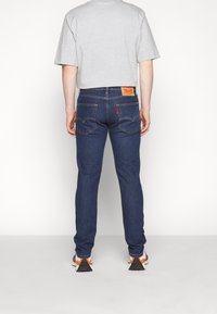 Modré denimové džíny s úzkým střihem, s žlutou koženou cedulkou na zadní straně, jemným prošíváním a volnočasovými botami s oranžovými akcenty.