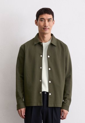 Olivgrüne Knopfjacke aus weichem Stoff, mit silbernen Druckknöpfen und klassischem Kragen, kombiniert mit einem hellen T-Shirt.