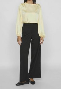 Nainen, jolla on päällään pitkähihaisen kermanvärisen paidan, mustat leveälahkeiset housut ja mustat ballerinat, seisoo yksinkertaista taustaa vasten.