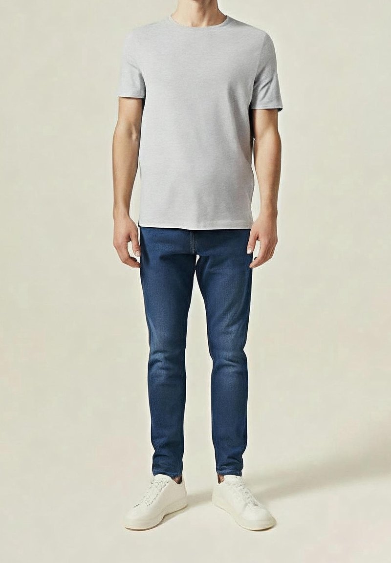 Homme portant un t-shirt gris clair uni, un jean bleu slim et des baskets blanches, debout devant un fond clair.