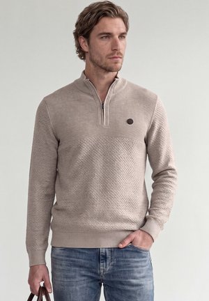 Mann trägt beigen strukturierten Viertel-Reißverschluss-Pullover und blaue Jeans, hält braunen Lederriemen in der linken Hand und schaut nach rechts vor schlichtem Hintergrund.