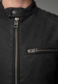 Chaqueta de cuero negro con un alto cuello, cremallera central plateada y un bolsillo adicional con cremallera, con un acabado texturizado en los hombros.