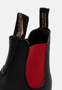 Stivale nero in pelle alla caviglia con pannelli elastici rossi ai lati. Presenta dettagli di cucitura e linguette con logo. Texture liscia e design resistente.