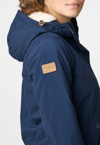 Granatowa kurtka z futrzanym kapturem, z brązową skórzaną naszywką z logo na rękawie oraz trwałą teksturą zewnętrznego materiału.