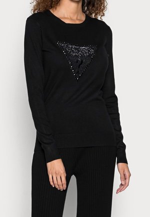 Mujer con suéter negro de manga larga con un logo triangular adornado que incluye un signo de interrogación en el pecho, combinado con pantalones negros.