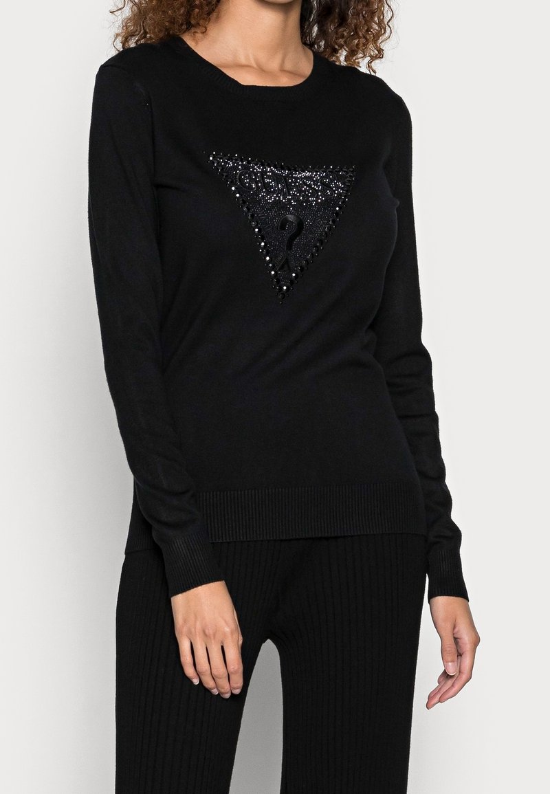Femme portant un pull à manches longues noir avec un logo triangulaire orné d'un point d'interrogation sur la poitrine, associé à un pantalon noir.