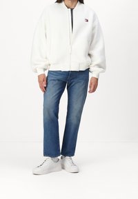 Man in een witte fleece zip-up jack, blauwe rechttoe rechtaan jeans, witte sokken en witte lage sneakers staande op een witte vloer.