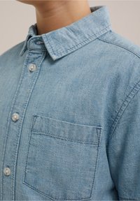 Světle modrá denimová košile s klasickým límečkem, zapínáním na knoflíky a náprsní kapsou s viditelným švy.