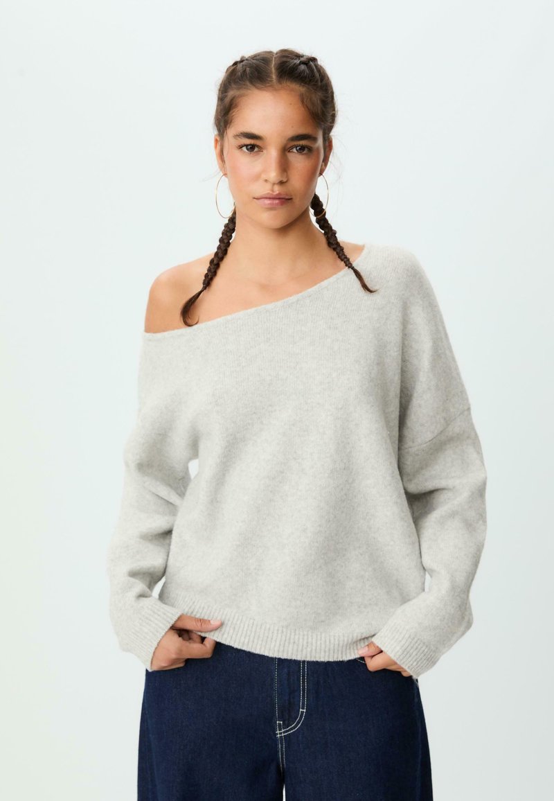 Hellgrauer, oversized Pullover mit einem schulterfreien Design, geripptem Saum und langen Ärmeln, kombiniert mit dunkelblauen Jeans. Weicher Stoff.