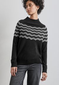 Schwarzer Pullover mit hohem Kragen, der ein Wellenmuster in Weiß über der Brust aufweist. Hergestellt aus einem weichen, strukturierten Stoff.