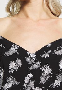 Kvinna med ljus hy och blont hår iklädd en svart off-shoulder-klänning med vita blommönster.