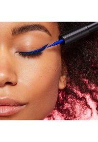 Eyeliner liquido blu applicato su un occhio chiuso con lunghe ciglia nere. La persona ha la pelle liscia e i capelli ricci, su uno sfondo rosa.