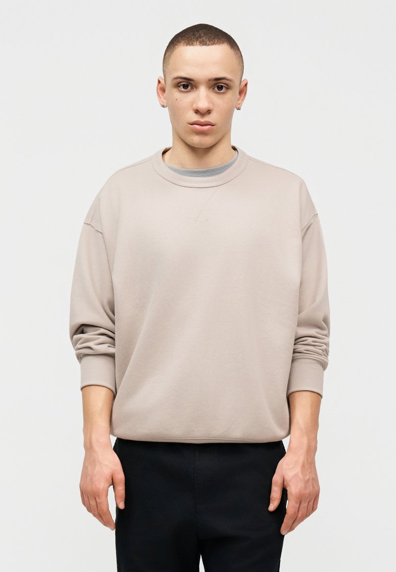 Giovane con capelli corti che indossa una felpa beige oversize e pantaloni neri, in piedi davanti a uno sfondo chiaro e uniforme.