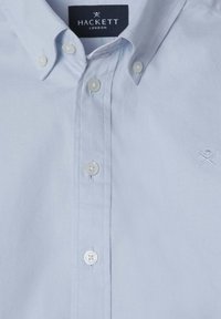 Světle modrá košile na knoflíky vyrobená z bavlny, s bílými knoflíky a vyšitým logem na hrudi. Hladká textilie, klasický design.