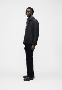 Casaco preto com fecho de botões, padrão texturizado na parte frontal, combinado com calças pretas e sapatos pretos, em pé contra um fundo branco.