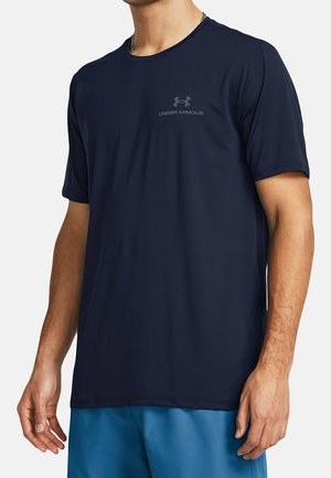Homme portant un T-shirt à manches courtes Under Armour bleu marine et un short de sport bleu, debout les bras détendus le long du corps.