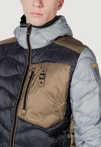 Polstret jakke med en blanding af sorte, khaki og grå materialer. Indeholder lynlåse foran, hætte og et logo-patch på ærmet.