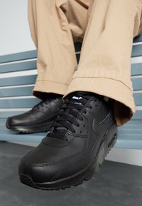 Person som bär beige byxor och svarta Nike Air Max sneakers sitter på en blå ribbad bänk med fötterna något upphöjda.