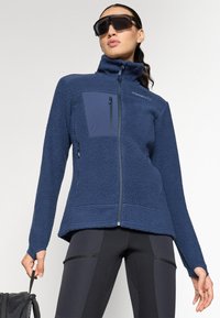 Námornícky modrá fleece bunda s vysokým golierom, zipsovými vreckami a textúrovanou látkou. Dizajn má priliehavý strih a kontrastný panel z inej látky.