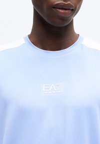 Κοντινό ενός ατόμου που φοράει ανοιχτό γαλάζιο πουκάμισο Emporio Armani EA7, εμφανίζοντας τον λαιμό, τα χείλη και το λογότυπο στο στήθος.