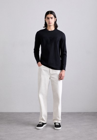 Melnas garām piedurknēm krekls ar mazu logotipu, baltas denima bikses un melni sporta apavi ar baltiem zoliņām, stāvot uz neitrāla fona.