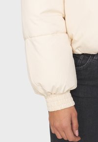 Giacca a imbottitura color crema con texture trapuntata, caratterizzata da polsini elasticizzati e una vestibilità comoda, abbinata a denim scuro.