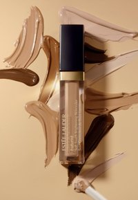 Estée Lauder Futurist Soft Touch Brightening Skincarer en un tubo transparente con acento dorado, rodeado de espirales de diferentes tonos de piel.