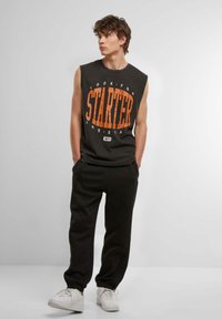 Black sleeveless shirt with orange graphic text "STARTER" and gray sweatpants. White sneakers; casual, sporty outfit.