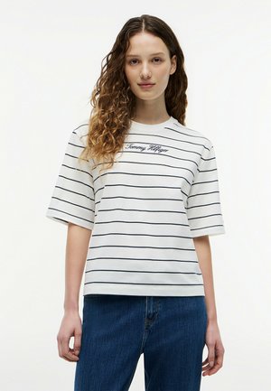 Młoda kobieta o kręconych włosach ubrana w białą, krótki rękaw, pasiastą koszulkę z napisem "Tommy Hilfiger" i niebieskie dżinsy, stojąca na białym tle.