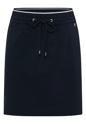 Kvinde iført hvid snørelukning hættetrøje, sort snørede nederdel og sølvfarvede sneakers, stående op ad en beige væg og kigger sidelæns.