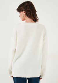 Pulover alb supradimensionat dintr-un material texturat și pufoasă; prezintă o guler larg și umeri lăsați, asortat cu blugi din denim bleumarin.