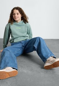 Felpa verde con cappuccio con logo "element", jeans blu a gamba larga e sneakers beige con suole in gomma, seduto su un pavimento grigio.