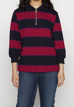 Vrouw die een poloshirt met lange mouwen draagt met brede horizontale rode en marineblauwe strepen en een blauwe spijkerbroek.