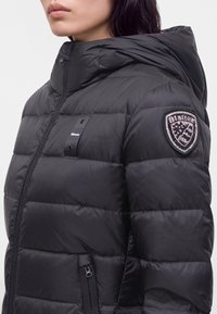 Femme portant une doudoune matelassée noire avec capuche et un patch de marque sur le haut du bras, fermeture éclair visible et poche avant.