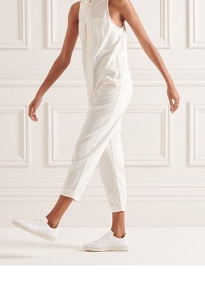 Personne marchant vêtue d'une combinaison sans manches blanche et de baskets blanches, devant un mur à panneaux.