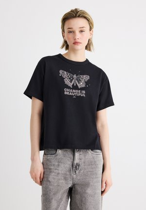 Jeune personne portant un t-shirt noir avec un motif de papillon et le texte « Le changement est beau », associé à un jean gris clair.