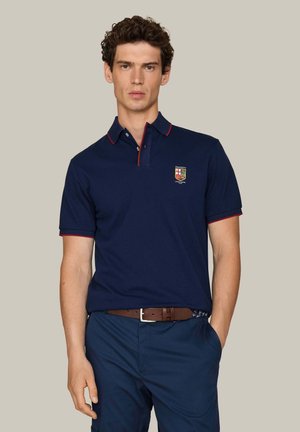 Jonge man die een marineblauw poloshirt met rode bies en een geborduurd wapen draagt, gecombineerd met marineblauwe broek en een bruine riem, staand tegen een beige achtergrond.